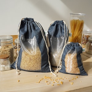 Puede incluir: Tres bolsas reutilizables de mezclilla con cordón de diferentes tamaños llenas de pasta, lentejas y guisantes partidos. Las bolsas tienen una ventana transparente y están dispuestas sobre una superficie de madera, con otros alimentos al fondo.