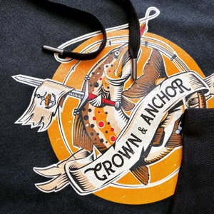 Puede incluir: Una sudadera con capucha negra con un gráfico circular amarillo que presenta una trucha marrón con una espada y una pancarta que dice "Crown & Anchor".