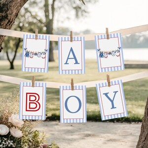 Può includere: Uno striscione con la scritta "A BOY" con bordi a righe blu e bianche e contorni rossi. Le lettere sono blu e rosse. Lo striscione è decorato con vestiti per bambini e tenuto da mollette di legno.