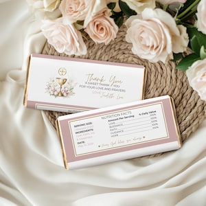 May include: Two chocolate bar wrappers with a pink and gold design. The top wrapper says "Thank You" with a floral and chalice graphic. The bottom wrapper has a nutrition facts label and the text "May God bless you always."