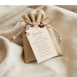 Może przedstawiać: Mały jutowy woreczek na prezent związany sznurkiem, z ozdobną etykietą. Na etykiecie napis "Thank You" z serdeczną wiadomością i kwiatowymi akcentami w kolorze bordowym i białym. Woreczek leży na neutralnej tkaninie.