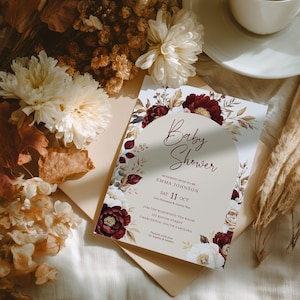 Może przedstawiać: Zaproszenie na baby shower z kwiatowym wzorem w kolorze bordowym, kremowym i beżowym. Tekst brzmi "Baby Shower" i zawiera datę, godzinę i miejsce. Zaproszenie otoczone jest suszonymi kwiatami i filiżanką kawy.
