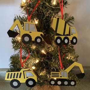 May include: Four wooden Christmas ornaments shaped like construction vehicles. The ornaments are yellow with black details and are hanging from a green Christmas tree.