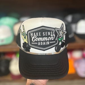May include: Black and white trucker hat with the text "MAKE SENSE COMMON AGAIN" on a white patch. The hat features a black brim, a gold rope detail, and patches of a rock on hand, a star, and cherries.