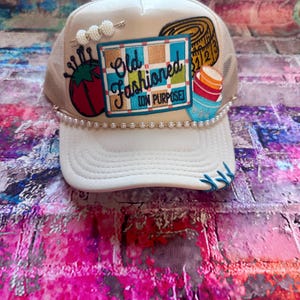 May include: A white trucker hat with a mesh back and a curved brim. The hat features a patch with the words "Old Fashioned (on purpose)" and sewing-themed embellishments, including a tomato, measuring tape, and a spool of thread. A pearl chain adorns the brim.