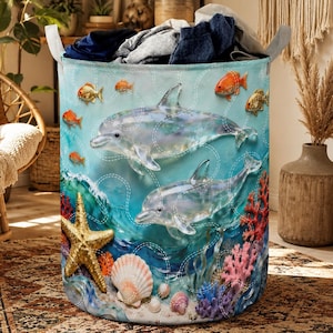 Könnte beinhalten: Ein zylindrischer Wäschekorb mit maritimem Thema. Das Design zeigt zwei Delfine, Seesterne, Muscheln und farbenfrohe Korallen vor einem blauen Ozeanhintergrund. Goldfische schwimmen um die Delfine. Der Korb hat weiße Griffe und ist mit Kleidung gefüllt.