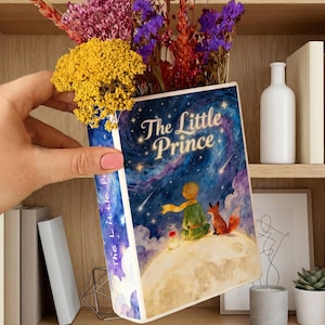May include: A book-shaped vase featuring the cover art of "The Little Prince" with a starry night sky, a boy, and a fox. The vase holds a bouquet of colorful dried flowers. The book's spine is visible, and the title is displayed on the front.
