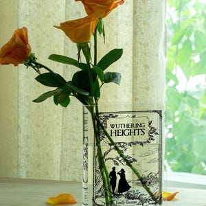 May include: A clear glass vase shaped like a book, featuring the cover of "Wuthering Heights" by Emily Bront&euml;. The vase holds orange roses with green stems and leaves. Scattered petals add color.