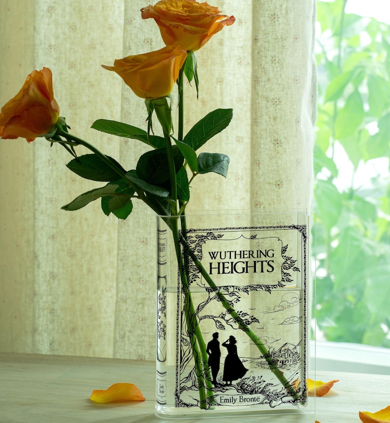 May include: A clear glass vase shaped like a book, featuring the cover of "Wuthering Heights" by Emily Bront&euml;. The vase holds orange roses with green stems and leaves. Scattered petals add color.