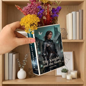 May include: A book titled "The Knight and the Moth" with a woman in armour on the cover, held in front of a wooden bookshelf. The book's spine is visible, and the title is also printed on it. The shelf is decorated with flowers.