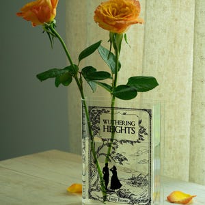 May include: A clear, rectangular vase with the cover of "Wuthering Heights" by Emily Bront&euml; printed on it. Two orange roses with green stems and leaves are in the vase. The vase sits on a light-colored wooden surface.