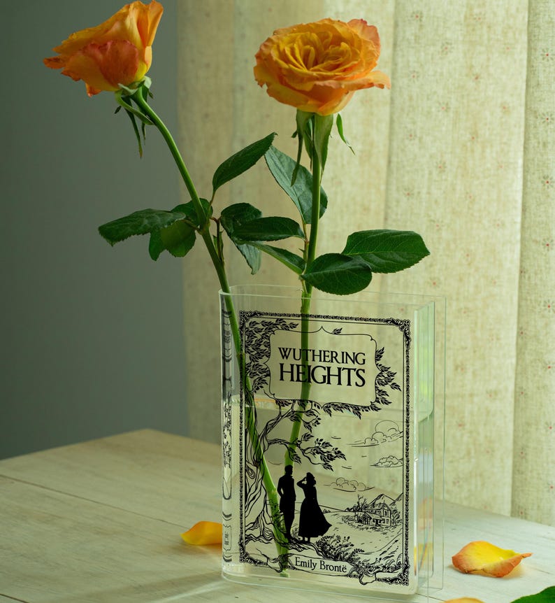 May include: A clear, rectangular vase with the cover of "Wuthering Heights" by Emily Bront&euml; printed on it. Two orange roses with green stems and leaves are in the vase. The vase sits on a light-colored wooden surface.