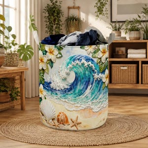 Könnte beinhalten: Ein zylindrischer Wäschekorb mit Strandmotiv. Das Kunstwerk zeigt eine große blaue und weiße Welle, Muscheln und Seesterne an einem Sandstrand. Weiße Blumen und grüne Blätter sind ebenfalls Teil des Designs. Der Korb ist mit gefalteter Kleidung gefüllt.