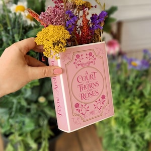 Könnte beinhalten: Eine rosa, buchförmige Vase mit dem Titel "A Court of Thorns and Roses" in goldfarbenen Buchstaben, gefüllt mit bunten Trockenblumen. Die Vase wird in einer Hand gehalten, mit einem verschwommenen Hintergrund aus Grün und Blumen.