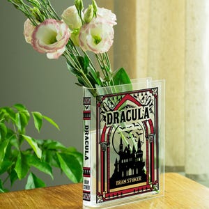 May include: A clear acrylic vase shaped like a book, featuring the title "DRACULA" and a gothic castle illustration. The vase holds white and pink flowers. The book's spine also displays the title and author's name.