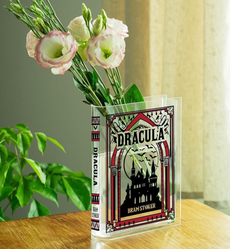 May include: A clear acrylic vase shaped like a book, featuring the title "DRACULA" and a gothic castle illustration. The vase holds white and pink flowers. The book's spine also displays the title and author's name.