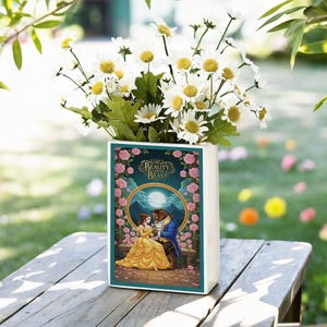 Può includere: Un vaso rettangolare bianco con un design di "La Bella e la Bestia", pieno di margherite bianche. Il vaso presenta uno sfondo turchese con un'immagine dorata di Belle e la Bestia. Il vaso è su un tavolo di legno.