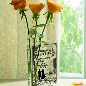 May include: Three orange roses in a clear vase featuring the cover art of "Wuthering Heights" by Emily Bront&euml;. The vase has a rectangular shape and is placed near a window with natural light.