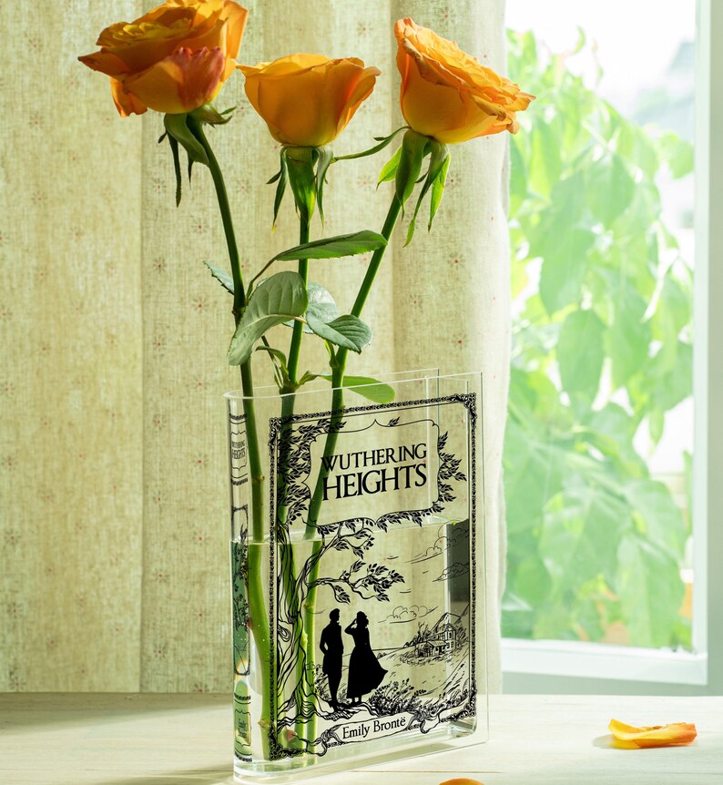 May include: Three orange roses in a clear vase featuring the cover art of "Wuthering Heights" by Emily Bront&euml;. The vase has a rectangular shape and is placed near a window with natural light.