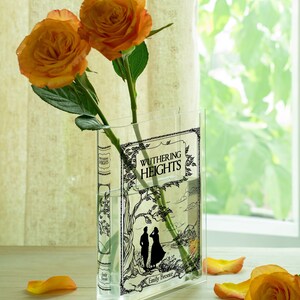 May include: A clear glass vase shaped like a book, featuring the title "Wuthering Heights" and an illustration. Two orange roses with green stems and leaves are in the vase. Scattered orange rose petals are on the surface.