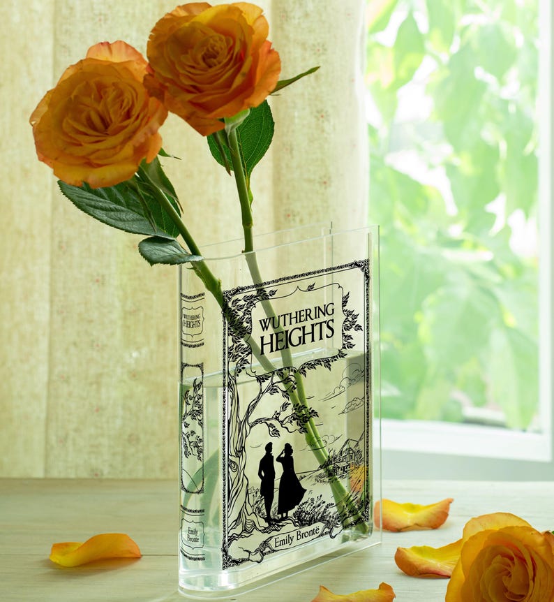 May include: A clear glass vase shaped like a book, featuring the title "Wuthering Heights" and an illustration. Two orange roses with green stems and leaves are in the vase. Scattered orange rose petals are on the surface.