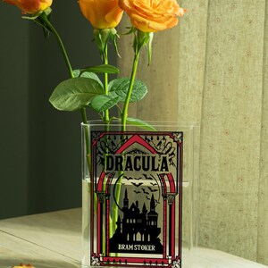 May include: A clear rectangular vase with the word "DRACULA" in black and red lettering, with a silhouette of a castle and bats. The vase holds orange roses. The author's name, Bram Stoker, is below the title.