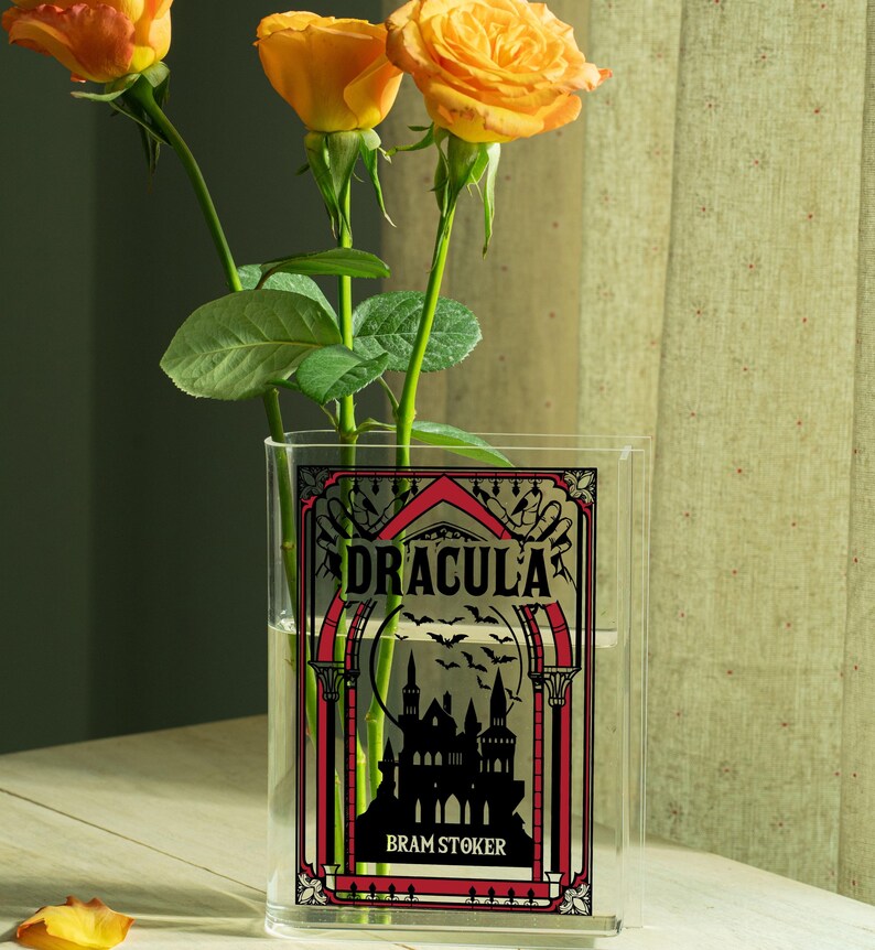 May include: A clear rectangular vase with the word "DRACULA" in black and red lettering, with a silhouette of a castle and bats. The vase holds orange roses. The author's name, Bram Stoker, is below the title.
