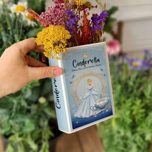 Könnte beinhalten: Eine hellblaue, buchförmige Vase mit dem Titel "Cinderella" und einer Illustration von Cinderella in einem blauen Kleid. Die Vase ist mit bunten Trockenblumen gefüllt. Der Text "When The Clock Strikes Twelve" ist ebenfalls sichtbar.