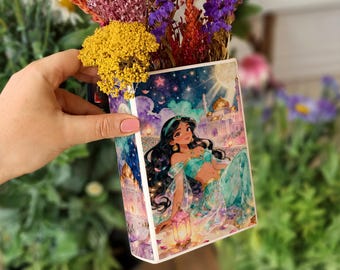 May include: A decorative floral container featuring a colorful illustration of Princess Jasmine from Aladdin. The container is filled with dried flowers in shades of yellow, purple, and red. The artwork depicts a palace scene.