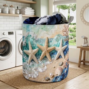 Könnte beinhalten: Ein zylindrischer Wäschekorb mit Strandmotiv. Der Korb zeigt eine Strandszene mit Meereswellen, Seesternen, Muscheln und Perlen. Die Farben umfassen Blau-, Beige- und Weißtöne. Der Korb ist mit gefalteter Kleidung gefüllt.