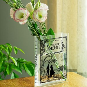 May include: A clear acrylic vase shaped like a book, featuring the cover art of "Wuthering Heights" by Emily Bront&euml;. The vase holds pink and white flowers with green stems and leaves. The vase is on a wooden surface.