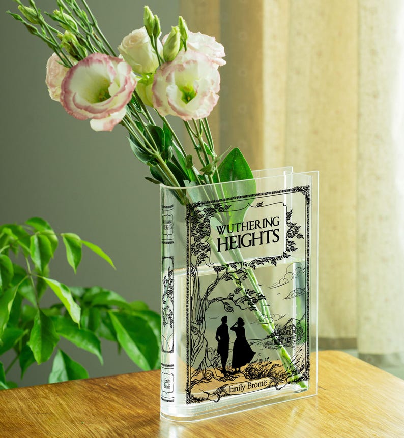 May include: A clear acrylic vase shaped like a book, featuring the cover art of "Wuthering Heights" by Emily Bront&euml;. The vase holds pink and white flowers with green stems and leaves. The vase is on a wooden surface.