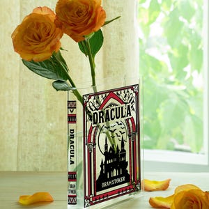 May include: A clear acrylic vase shaped like a book, featuring the cover of "Dracula" by Bram Stoker. The vase holds two orange roses. The book cover design is black, red, and white, with a castle silhouette and bats.