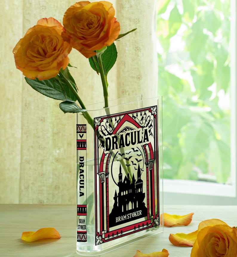 May include: A clear acrylic vase shaped like a book, featuring the cover of "Dracula" by Bram Stoker. The vase holds two orange roses. The book cover design is black, red, and white, with a castle silhouette and bats.
