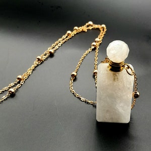 Pode incluir: Um pingente retangular de pedra clara com um tampão redondo facetado, suspenso por uma delicada corrente dourada. A corrente apresenta pequenas contas douradas espaçadas uniformemente. O fundo é de cor escura e sólida.