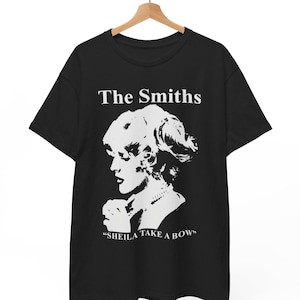 Puede incluir: Una camiseta negra de manga corta con un estampado gráfico blanco de una persona de perfil. El texto "The Smiths" aparece sobre la imagen y el texto "SHEILA TAKE A BOW" está impreso debajo del gráfico. La camiseta cuelga de una percha de madera.