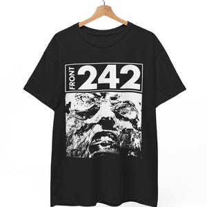 May include: Black t-shirt with the text "FRONT 242" above a black and white face graphic. The t-shirt is displayed on a wooden hanger suspended from a metal rail.