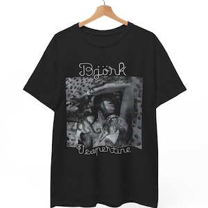 以下が含まれることがあります： 黒いTシャツ。人物のグレースケール画像と、画像の上に筆記体フォントで「Bjork」という文字がプリントされています。「Vespertine」という文字が画像の下に筆記体フォントでプリントされています。