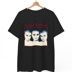 以下が含まれることがあります： 黒いTシャツ。赤色の長方形に囲まれた、髪を剃った3つの顔のグラフィック。画像の上には「Sinéad O'Connor」の文字が赤色で書かれています。Tシャツは木製のハンガーに掛けられています。