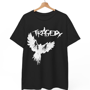 Op de afbeelding: Zwart T-shirt met het woord "TRAGEDY" in het wit, boven een witte afbeelding van een vogel met uitgespreide vleugels. Het T-shirt hangt aan een houten hanger.
