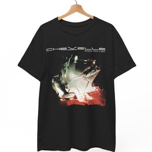 Puede incluir: Camiseta negra con el nombre de la banda "CHEVELLE" sobre un gráfico de un vaso roto con agua y detalles en rojo. El texto "WONDER WHAT'S NEXT" está debajo del nombre de la banda.
