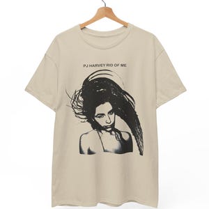Pode incluir: Camiseta bege com um gráfico a preto e branco de uma mulher com cabelo comprido. O texto "PJ HARVEY RID OF ME" está impresso acima do gráfico. A camisa está pendurada num cabide de madeira.