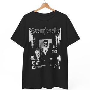 May include: Black t-shirt featuring the band name "Brujeria" in a gothic font. The graphic is a black and white image of a person with bottles and other objects. The shirt is hanging on a wooden hanger.