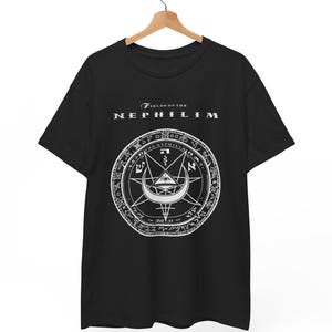 Puede incluir: Camiseta negra con un diseño gráfico blanco. El diseño incluye el texto "FIELDS OF THE NEPHILIM" sobre un emblema circular con símbolos y texto. La camiseta está colgada en una percha de madera.