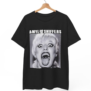 Puede incluir: Camiseta negra con una imagen en blanco y negro de una mujer con la boca abierta y la lengua fuera. El texto "AMYL AND THE SNIFFERS" está impreso encima de la imagen en letras blancas. La camiseta está colgada de una percha de madera.