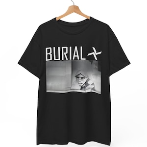 Op de afbeelding: Zwart T-shirt met het woord "BURIAL" in witte blokletters en een afbeelding. Onder de tekst staat een zwart-wit afbeelding van een persoon in een kledingstuk met capuchon. Het T-shirt hangt aan een houten hanger.