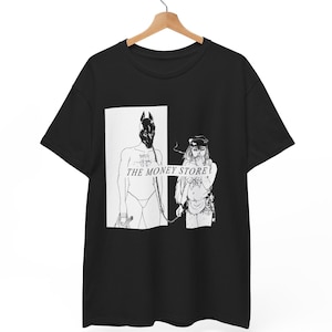 Puede incluir: Camiseta negra con un diseño gráfico que presenta dos figuras en una ilustración en blanco y negro. El texto "THE MONEY STORE" está impreso en la camiseta. La camiseta está colgada en una percha de madera.