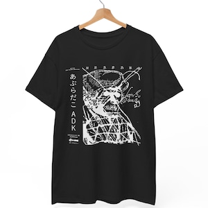 Puede incluir: Camiseta negra con un diseño gráfico blanco que presenta una cara estilizada y texto japonés. El diseño incluye el texto "あぶらだこ ADK" y otros caracteres japoneses. La camiseta está colgada en una percha de madera.