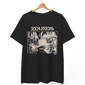 May include: Black t-shirt with a graphic design featuring the word "Zounds" in a stylized font. The design includes an illustration and text that reads "To govern me is a usurper and a tyrant." The shirt is hanging on a wooden hanger.