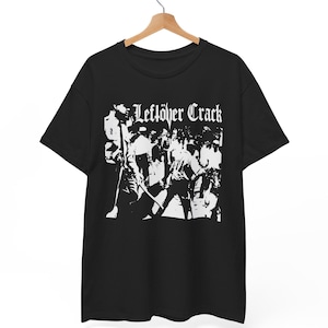 May include: Black t-shirt with the white text "Leftöver Crack" above a white graphic of a crowd of people. The shirt is hanging on a wooden hanger against a white background.